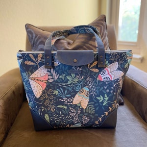 May include: A dark blue tote bag with a floral and insect print. The bag has a zipper closure and two leather handles. The bag is sitting on a brown couch.