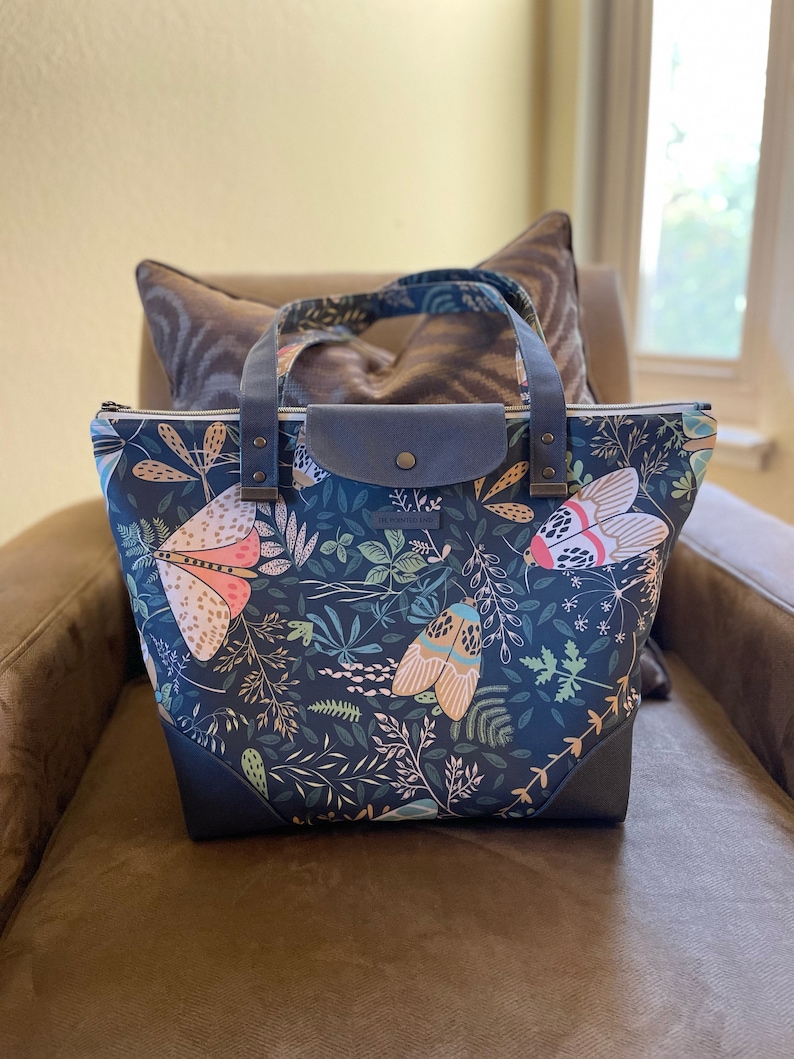 May include: A dark blue tote bag with a floral and insect print. The bag has a zipper closure and two leather handles. The bag is sitting on a brown couch.