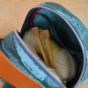 May include: A teal and green patterned zippered bag with a ball of white yarn and two wooden knitting needles inside. The bag has a brown leather strap.