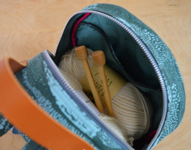 May include: A teal and green patterned zippered bag with a ball of white yarn and two wooden knitting needles inside. The bag has a brown leather strap.