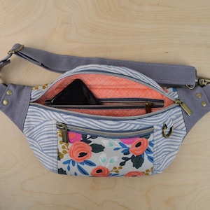 May include: A gray and white patterned fanny pack with a zippered pocket and a floral print. The fanny pack has a gray adjustable strap and a brass buckle.