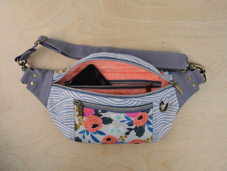 May include: A gray and white patterned fanny pack with a zippered pocket and a floral print. The fanny pack has a gray adjustable strap and a brass buckle.