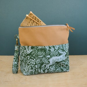 May include: A green and tan fabric project bag with a zipper closure and a white printed design of rabbits and flowers. The bag has a brown leather strap and a brown leather zipper pull.