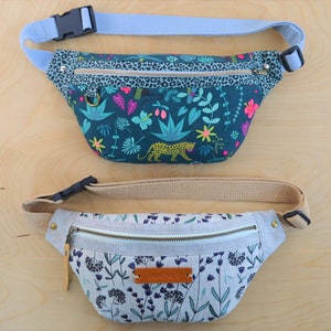 May include: Two fanny packs with adjustable straps. The top fanny pack has a green, blue, and pink floral print with a leopard. The bottom fanny pack has a light gray background with a floral print in shades of blue and green. The bottom fanny pack has a brown leather tag with the word "remarkable" printed on it.