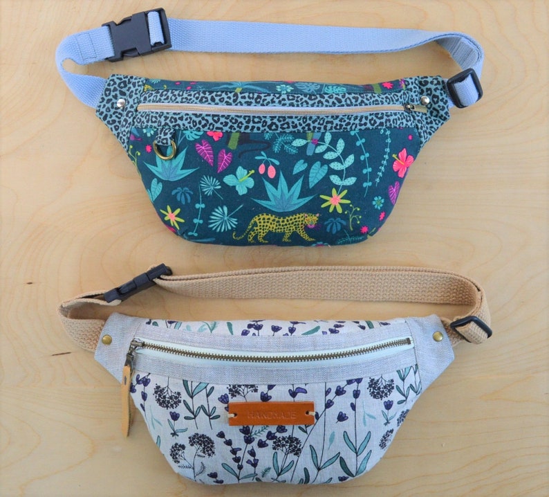 May include: Two fanny packs with adjustable straps. The top fanny pack has a green, blue, and pink floral print with a leopard. The bottom fanny pack has a light gray background with a floral print in shades of blue and green. The bottom fanny pack has a brown leather tag with the word "remarkable" printed on it.