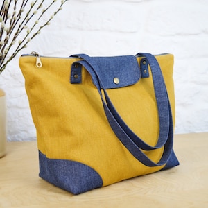 May include: A yellow tote bag with blue denim accents. The bag has two long straps and a zipper closure.