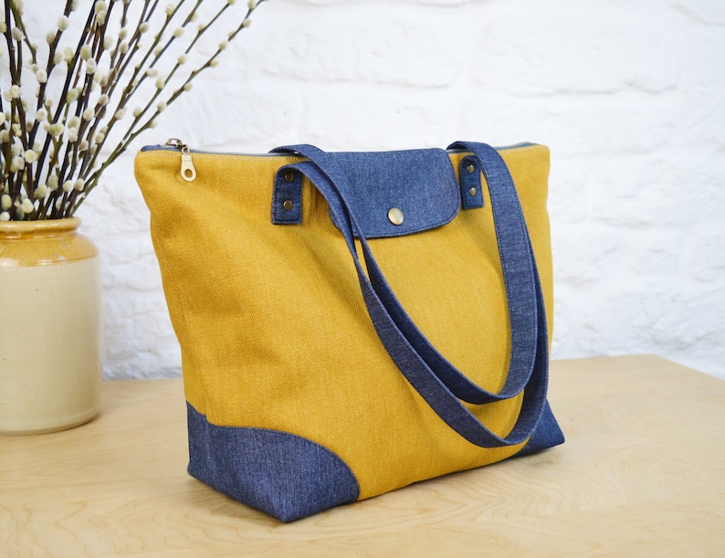 May include: A yellow tote bag with blue denim accents. The bag has two long straps and a zipper closure.
