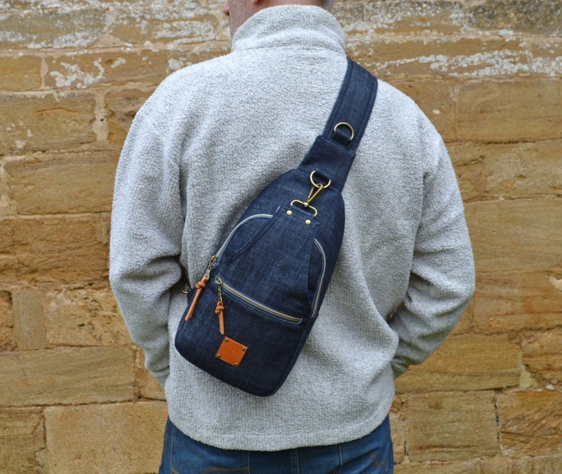 May include: A denim crossbody bag with a brown leather strap and a zippered pocket. The bag is worn by a person wearing a gray sweater and blue jeans.