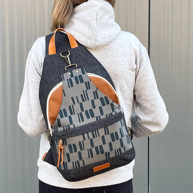 May include: A gray and black canvas backpack with a brown leather flap and a geometric pattern. The backpack has a single adjustable strap and a zipper closure.