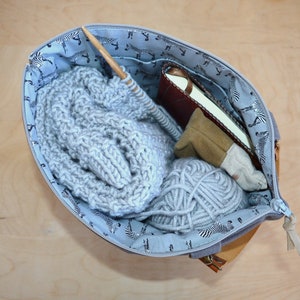May include: A light blue zippered tote bag with a zebra print lining. The bag is open and filled with a gray knitted scarf, a ball of gray yarn, a brown leather journal, and a small brown pouch.