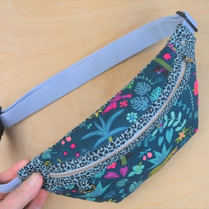 May include: A blue and green fanny pack with a zipper closure and a leopard print trim. The fanny pack has a tropical print with palm trees, flowers, and birds.