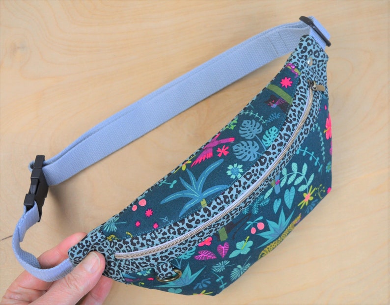 May include: A blue and green fanny pack with a zipper closure and a leopard print trim. The fanny pack has a tropical print with palm trees, flowers, and birds.