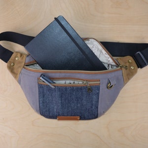 May include: A gray canvas fanny pack with a brown leather flap and a denim pocket. The fanny pack has a black zipper and a black strap. A black notebook and a pen are inside the fanny pack.