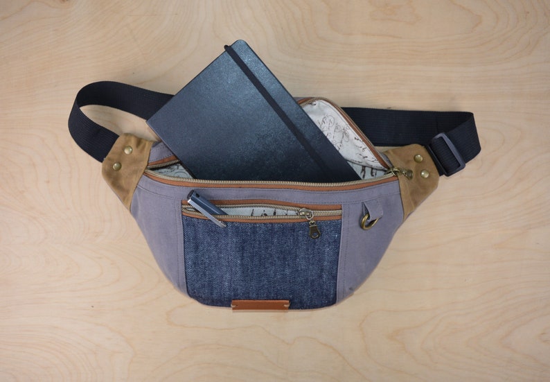 May include: A gray canvas fanny pack with a brown leather flap and a denim pocket. The fanny pack has a black zipper and a black strap. A black notebook and a pen are inside the fanny pack.