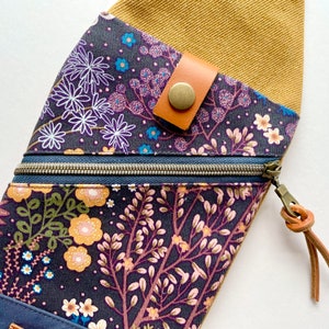 K&ouml;nnte beinhalten: Eine kleine Rei&szlig;verschlusstasche mit Blumenmuster und einem braunen Lederriemen mit einem Metallknopf. Die Tasche ist aus dunkelblauem Stoff mit einem Blumenmuster in den Farben Rosa, Gelb und Gr&uuml;n. Der Riemen ist oben an der Tasche befestigt und hat einen Metallknopf, mit dem der Riemen befestigt werden kann.