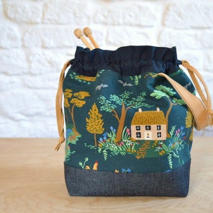 May include: A green drawstring bag with a cottage and trees print. The bag has a brown leather strap and a denim bottom.