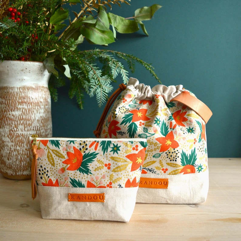 Pu&ograve; includere: Due borse in tessuto con stampa floreale con fiori rossi, foglie verdi e dettagli dorati. Le borse hanno uno sfondo crema e una tracolla in pelle marrone. La borsa pi&ugrave; grande ha la scritta "KANDOU" stampata sul fondo. La borsa pi&ugrave; piccola ha una cerniera e la scritta "KANDOU" stampata sul fondo.