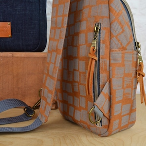 May include: A small orange and gray patterned backpack with a blue strap and brown leather accents. The backpack has two zippered pockets and a small leather tag that says "Mamboo Street".