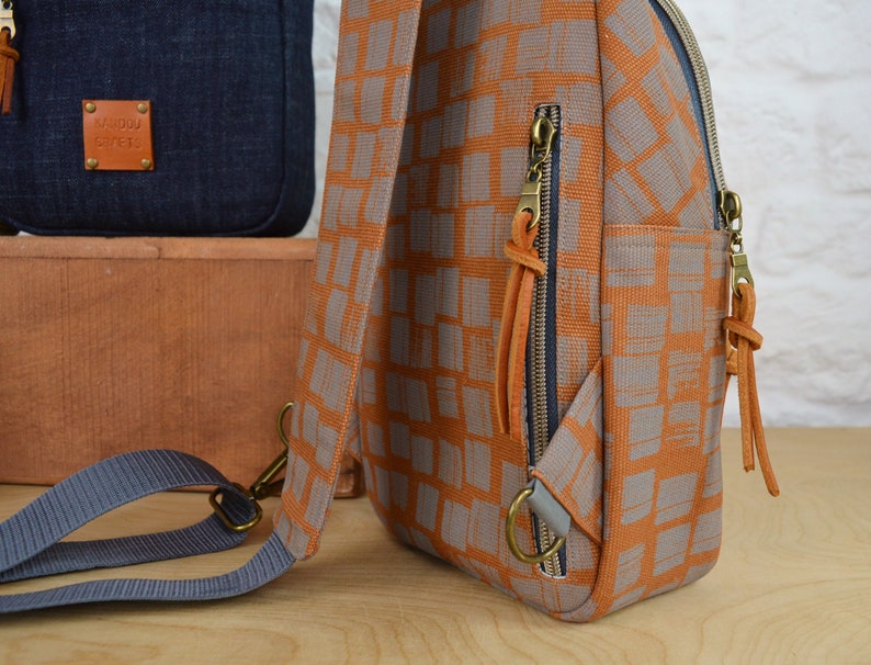 May include: A small orange and gray patterned backpack with a blue strap and brown leather accents. The backpack has two zippered pockets and a small leather tag that says "Mamboo Street".