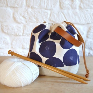 May include: A white yarn ball with a blue and white fabric drawstring bag with a brown leather handle. Two wooden knitting needles are next to the yarn ball.