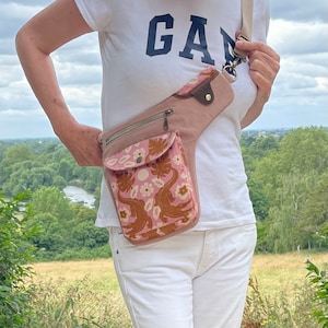 Pu&ograve; includere: Una marsupio in tela rosa con stampa floreale e un accento in pelle marrone. La borsa &egrave; indossata sul fianco di una persona che indossa una camicia bianca con la scritta "GAP" stampata su di essa.