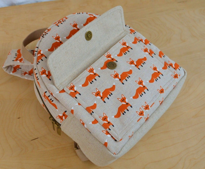 May include: A small, round, beige canvas backpack with a zip closure and a flap with a button closure. The backpack has a repeating pattern of orange foxes on a beige background.