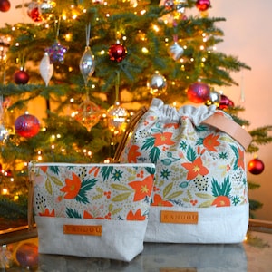 May include: Two fabric bags with a floral print featuring red poinsettia flowers, green leaves, and gold accents. The bags have a drawstring closure and a leather handle. The larger bag has the word "KANDOU" printed on the front. Both bags are sitting in front of a Christmas tree with lights.