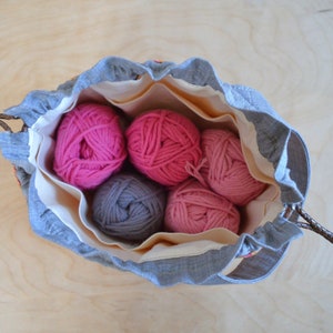 May include: A gray drawstring bag with a white lining, filled with five balls of yarn. Two balls are pink, two are a darker pink, and one is gray.