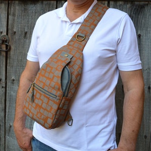 May include: A brown and gray patterned crossbody bag with a zipper closure and a gray pocket on the front. The bag is worn over the shoulder of a person wearing a white shirt.
