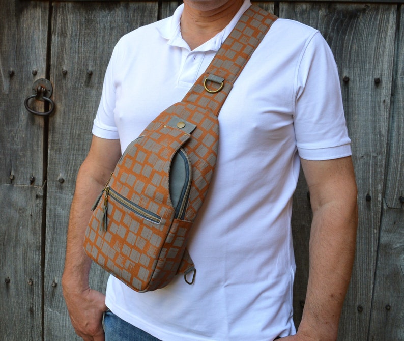 May include: A brown and gray patterned crossbody bag with a zipper closure and a gray pocket on the front. The bag is worn over the shoulder of a person wearing a white shirt.