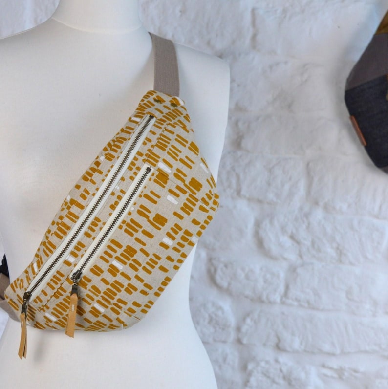 May include: A mustard yellow fanny pack with a white geometric pattern. The bag has two zipper closures and a long adjustable strap.