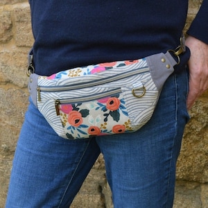 May include: A gray and white fabric fanny pack with a floral print and two zippered pockets. The fanny pack is worn around the waist of a person wearing blue jeans.