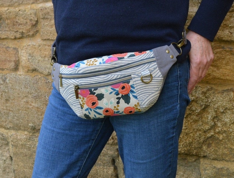 May include: A gray and white fabric fanny pack with a floral print and two zippered pockets. The fanny pack is worn around the waist of a person wearing blue jeans.