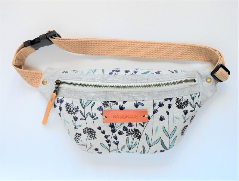 May include: A light gray fanny pack with a floral print featuring blue and green flowers. The fanny pack has a zipper closure and a brown leather strap with the word "HANDMADE" embossed on it.