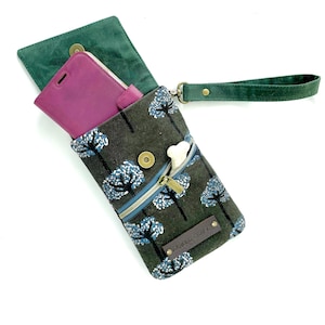 Puede incluir: Una funda para tel&eacute;fono con estampado verde oscuro y gris con cremallera y correa para la mu&ntilde;eca. La funda tiene un dise&ntilde;o de &aacute;rbol con detalles en azul y blanco. Se ve una funda de tel&eacute;fono morada dentro del compartimento principal. La etiqueta dice "NANDU CRAFT".