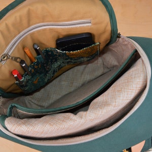 May include: A green canvas backpack with a zippered top opening. The inside of the backpack is lined with a beige fabric and has a zippered pocket. The pocket is filled with pens, pencils, and a phone.
