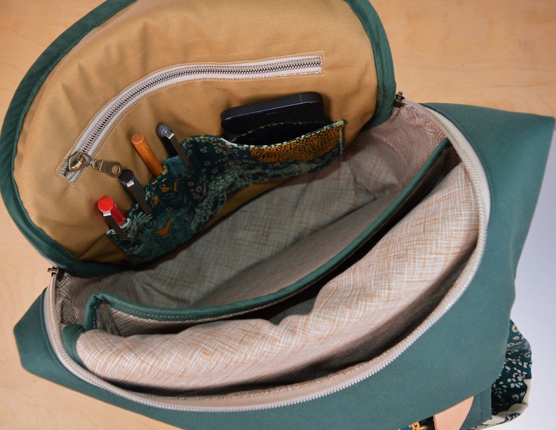 May include: A green canvas backpack with a zippered top opening. The inside of the backpack is lined with a beige fabric and has a zippered pocket. The pocket is filled with pens, pencils, and a phone.