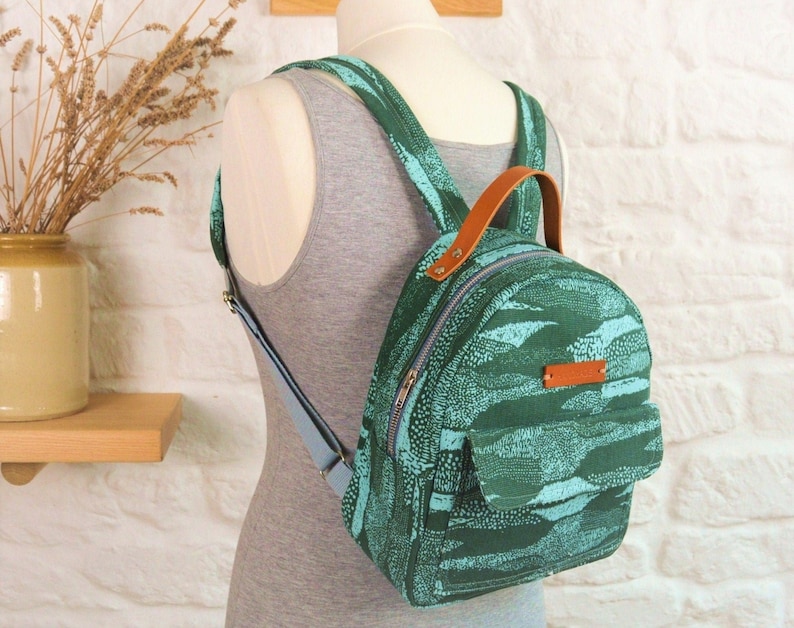 May include: A small green and blue patterned backpack with a brown leather handle and adjustable straps. The backpack has a zipper closure and a front pocket.