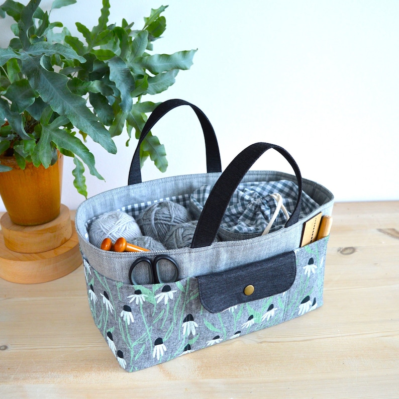 May include: A gray fabric tote bag with black handles, filled with gray yarn, knitting needles, and scissors. The bag features a floral pattern with white flowers and green stems. A small pocket with a button closure is on the front.