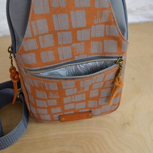 May include: A small, orange and gray canvas backpack with a zippered pocket and a brown leather strap. The backpack has a geometric pattern of squares.