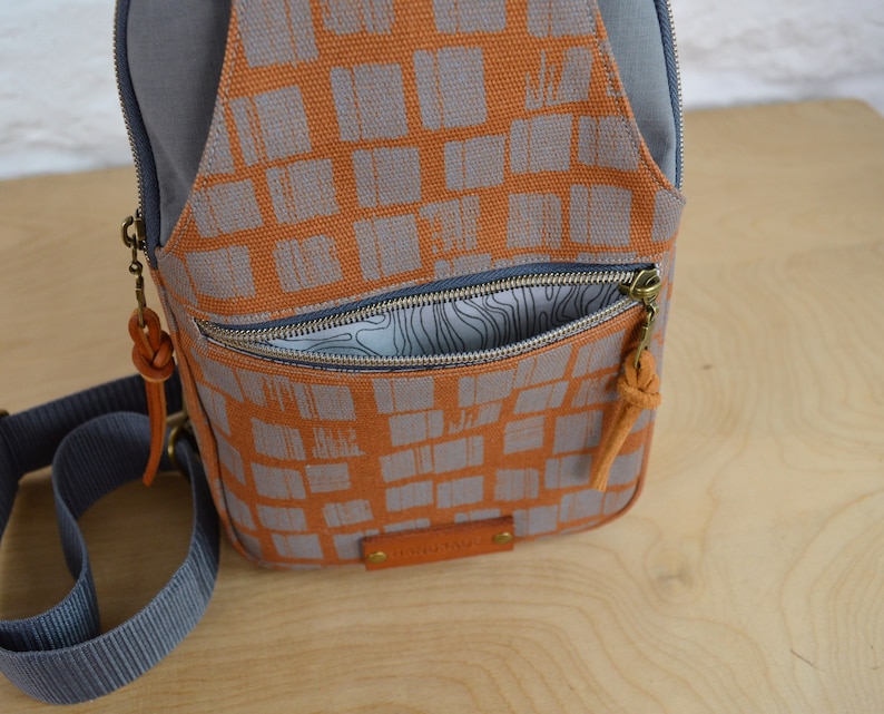 May include: A small, orange and gray canvas backpack with a zippered pocket and a brown leather strap. The backpack has a geometric pattern of squares.