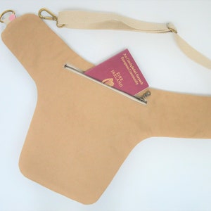 Pu&ograve; includere: Una borsa a tracolla in tela beige con una tasca con cerniera e una tracolla regolabile. La borsa &egrave; mostrata con un passaporto all'interno della tasca.