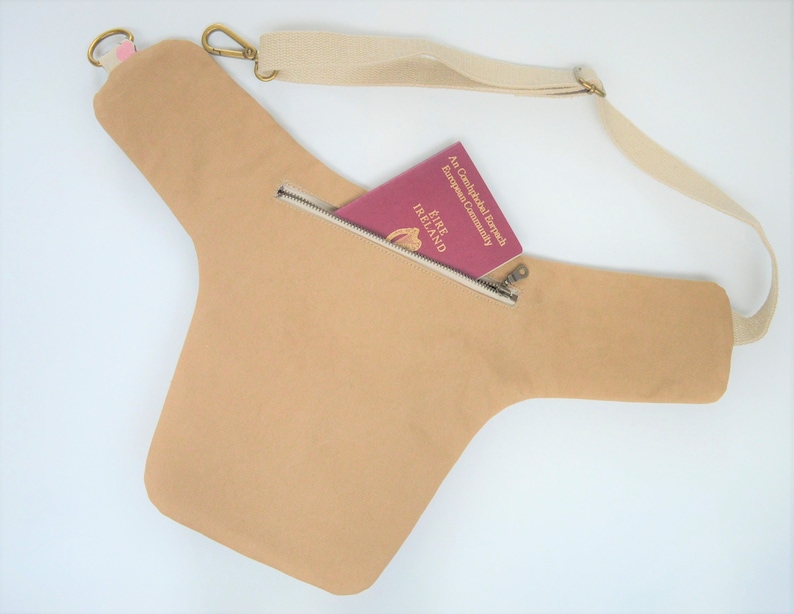 Pu&ograve; includere: Una borsa a tracolla in tela beige con una tasca con cerniera e una tracolla regolabile. La borsa &egrave; mostrata con un passaporto all'interno della tasca.
