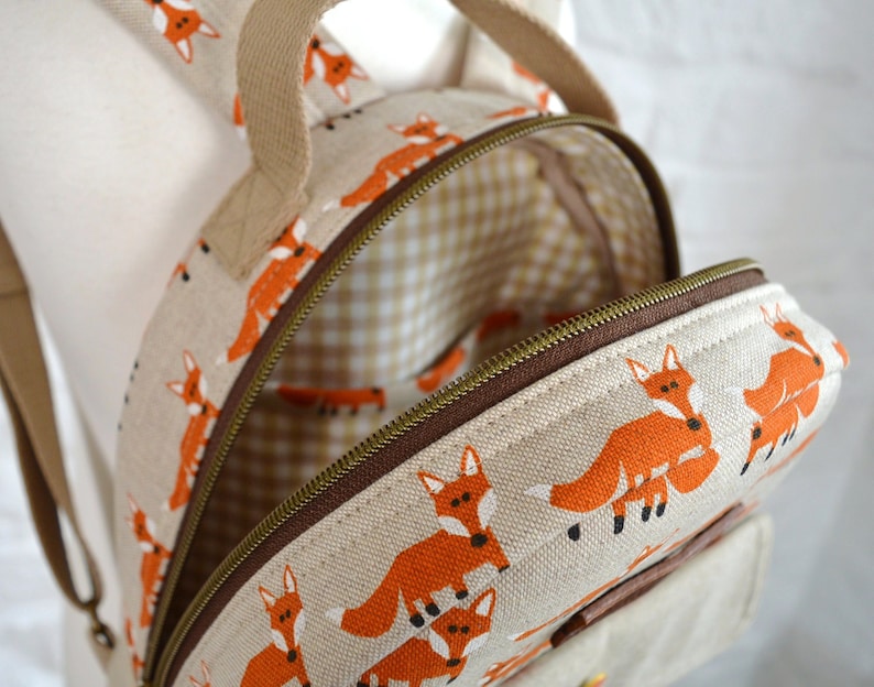 May include: A small beige canvas backpack with a zip closure. The backpack is decorated with a repeating pattern of orange foxes. The inside of the backpack is lined with a brown and white checked fabric.