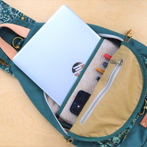 May include: A green backpack with a floral pattern and a tan zippered pocket. The backpack is open and a laptop, phone, and pens are visible inside.