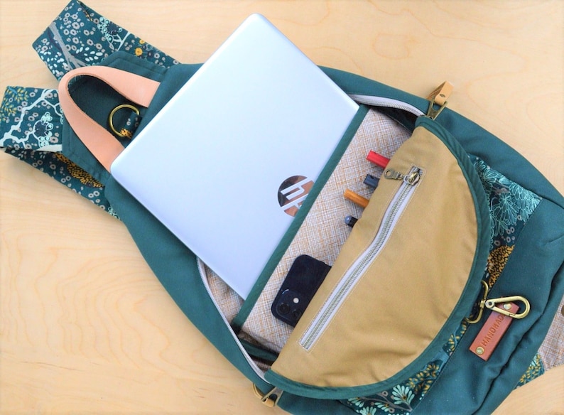 May include: A green backpack with a floral pattern and a tan zippered pocket. The backpack is open and a laptop, phone, and pens are visible inside.
