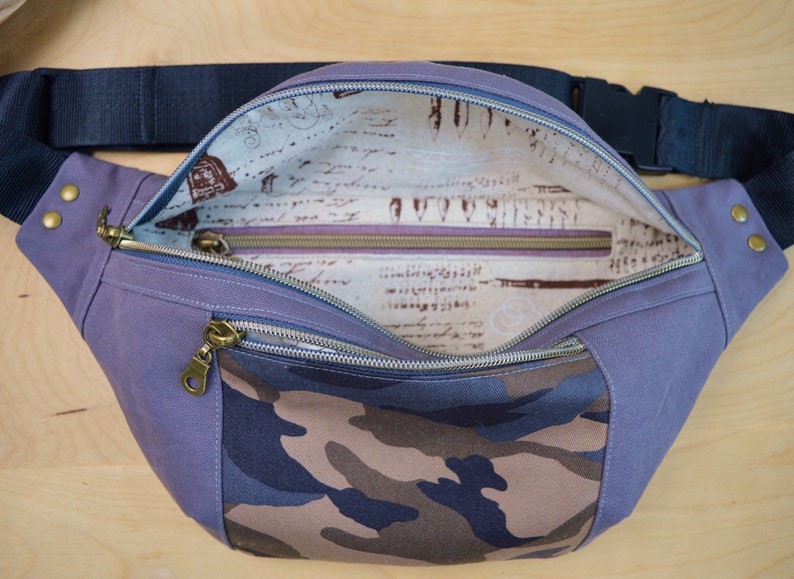 May include: A purple and camouflage fanny pack with a zippered pocket and a zippered closure. The inside of the bag is lined with a cream fabric with a brown and black print.