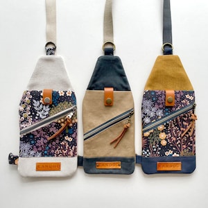 K&ouml;nnte beinhalten: Drei Umh&auml;ngetaschen mit Blumenmustern. Die Taschen sind aus Canvas und haben einen Rei&szlig;verschluss. Die Taschen sind in verschiedenen Farben: beige, schwarz und marineblau. Jede Tasche hat einen Lederriemen und eine Metallschnalle. Die Taschen haben den Schriftzug "KANDOU" aufgedruckt.