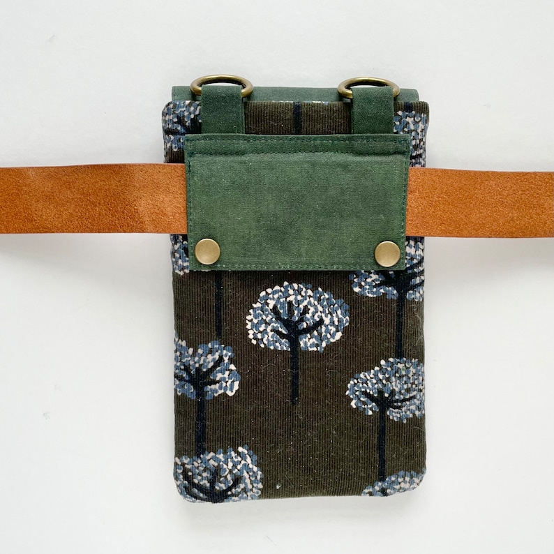 Puede incluir: Peque&ntilde;o bolso rectangular con un estampado de &aacute;rboles marr&oacute;n y azul, una solapa verde y un cintur&oacute;n de cuero marr&oacute;n. El bolso tiene dos anillos color lat&oacute;n en la parte superior y se asegura con dos broches color lat&oacute;n.