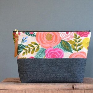 Pu&ograve; includere: Una pochette con zip a stampa floreale con fondo in denim nero. La pochette presenta una variet&agrave; di fiori rosa, rossi, gialli e verdi su uno sfondo bianco.